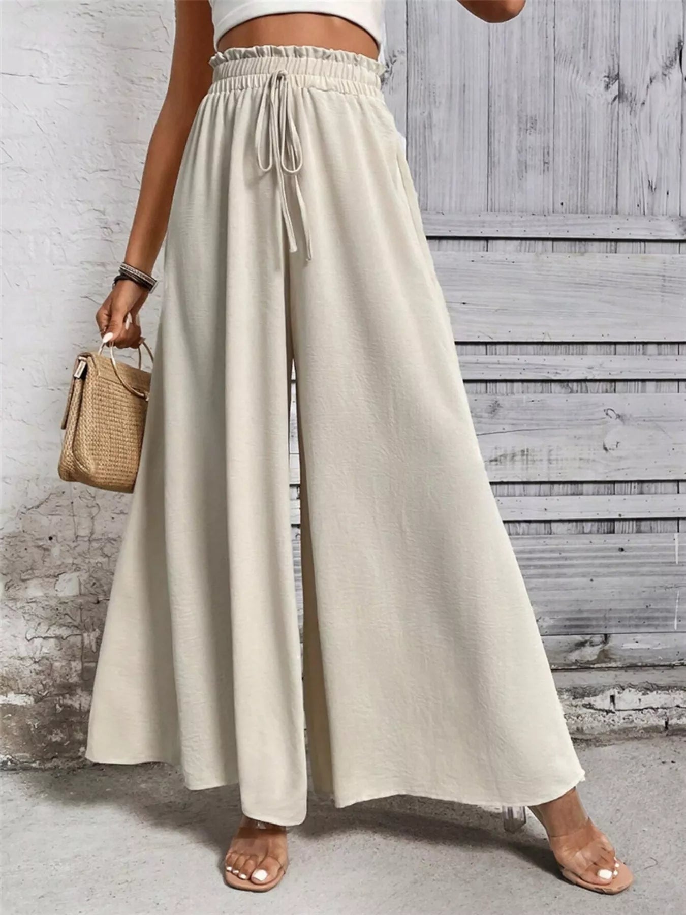 NEWYORKLISTED STYLE COLLECTION: Person wearing beige wide-leg pants with a white top against a wooden wall. 