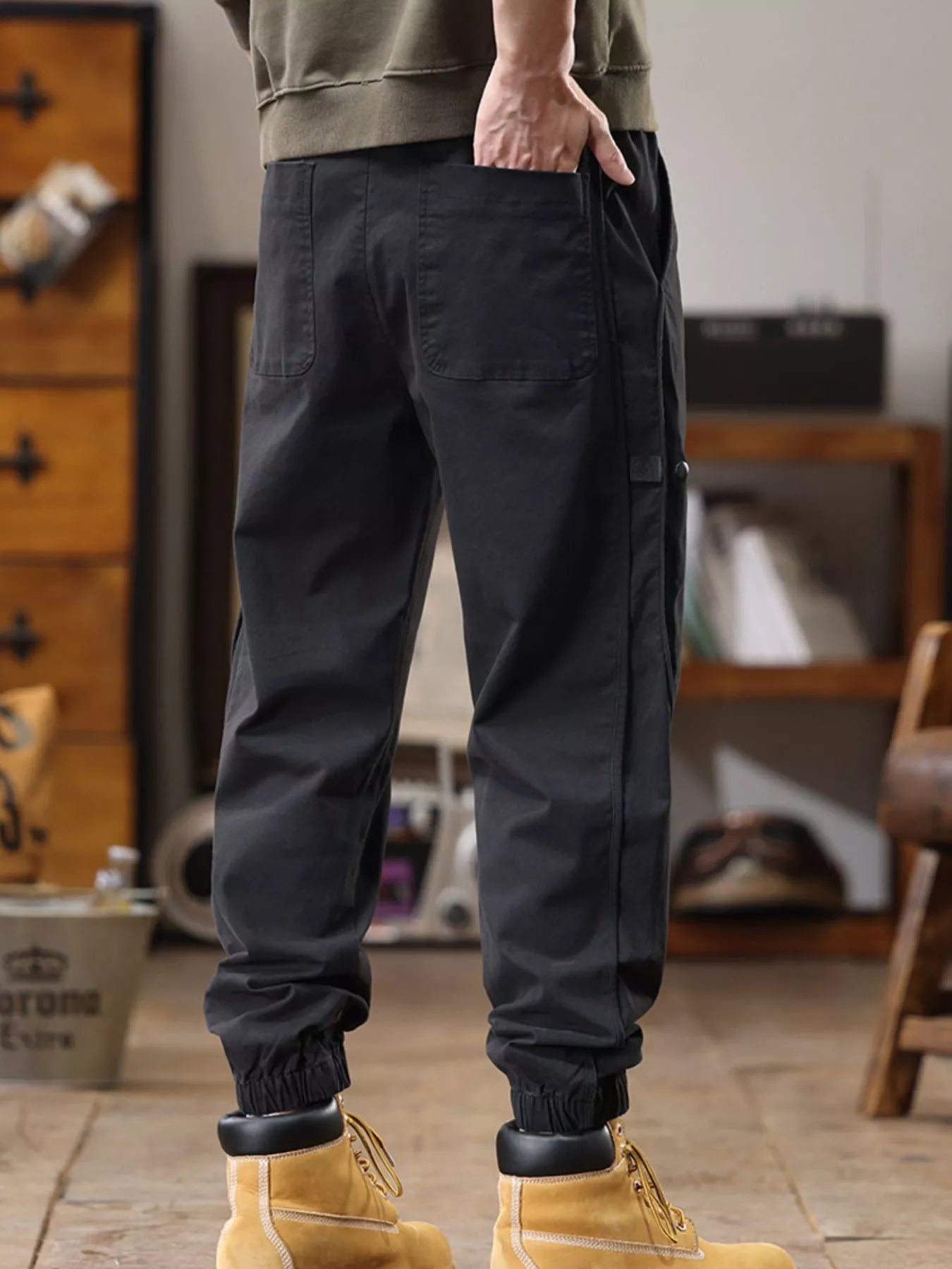NEWYORKLISTED ATLAS MEN'S COLLECTION: Person wearing dark cargo pants and yellow boots in a room with wooden furniture.