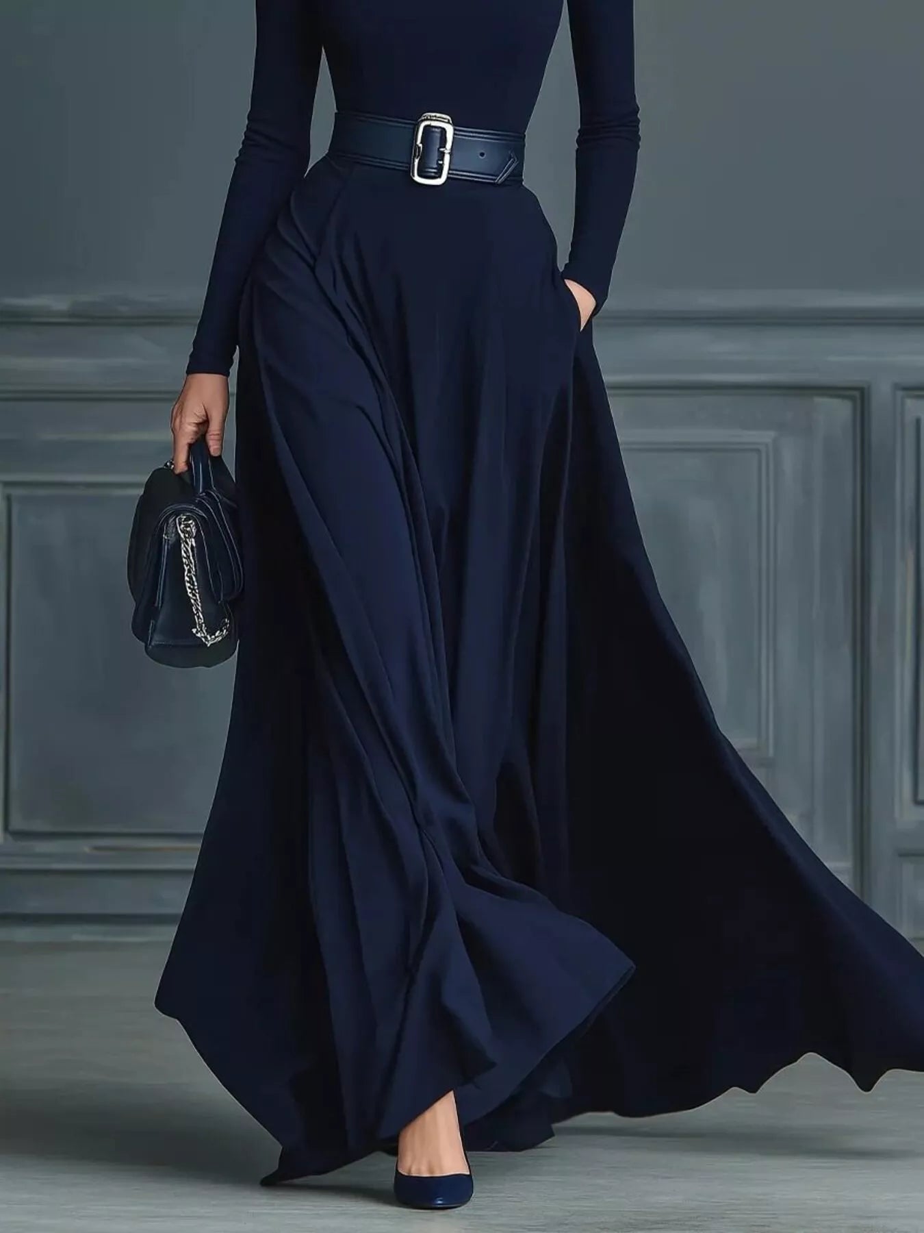 NEWYORKLISTED STYLE COLLECTION: Person wearing a long navy dress with a belt against a gray paneled wall.