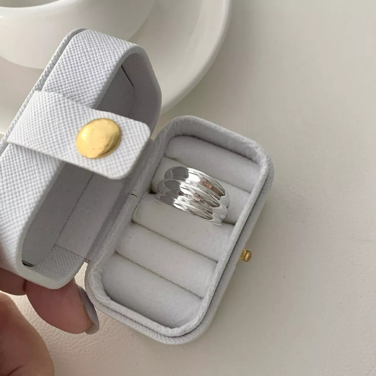 NYLD Glamour Collection: Silver ring in a white jewelry case with a gold button on a light background