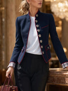 NEWYORKLISTED STYLE COLLECTION:  A woman models a navy blue military-style blazer with red piping and silver buttons, worn over a white top and paired with dark trousers. She is holding a burgundy handbag in a luxurious indoor setting with warm lighting.