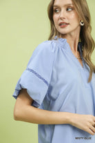 NEWYORKLISTED STYLE COLLECTION: Woman wearing a light blue blouse with embroidered sleeves against a green background