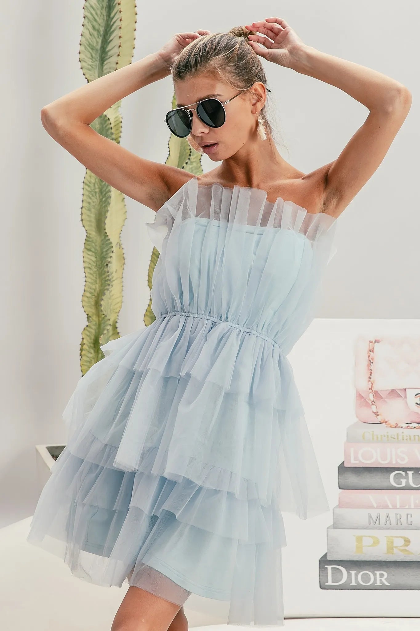 NEWYORKLISTED STYLE COLLECTION: Sleeveless Layered Organza Dress: Woman wearing a light blue off-shoulder dress with sunglasses, standing against a neutral background with books and a plant.