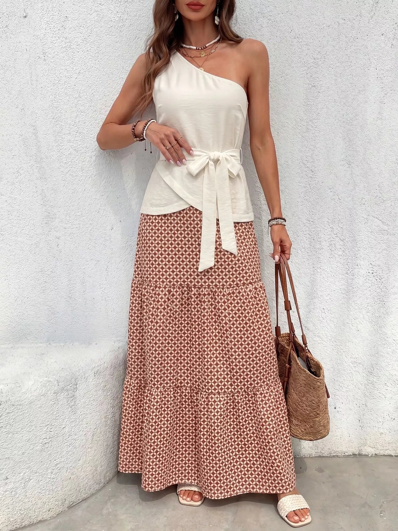 NEWYORKLISTED STYLE COLLECTION: Woman wearing a white one-shoulder top and patterned skirt with a brown bag against a light gray wall.