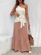 NEWYORKLISTED STYLE COLLECTION: Woman wearing a white one-shoulder top and patterned skirt with a brown bag against a light gray wall.
