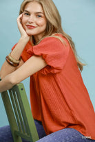 NEWYORKLISTED STYLE COLLECTION: Woman wearing a coral shirt sitting on a green chair against a light blue background