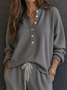NEWYORKLISTED STYLE & COMFORT COLLECTION: A woman wearing a gray waffle-knit long-sleeve henley top with white buttons and matching gray drawstring pants, standing against a dark textured wall.