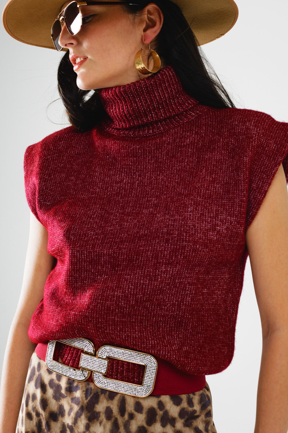 Woman wearing a red sleeveless knitted top with a belt, sunglasses, and a hat on a light gray background