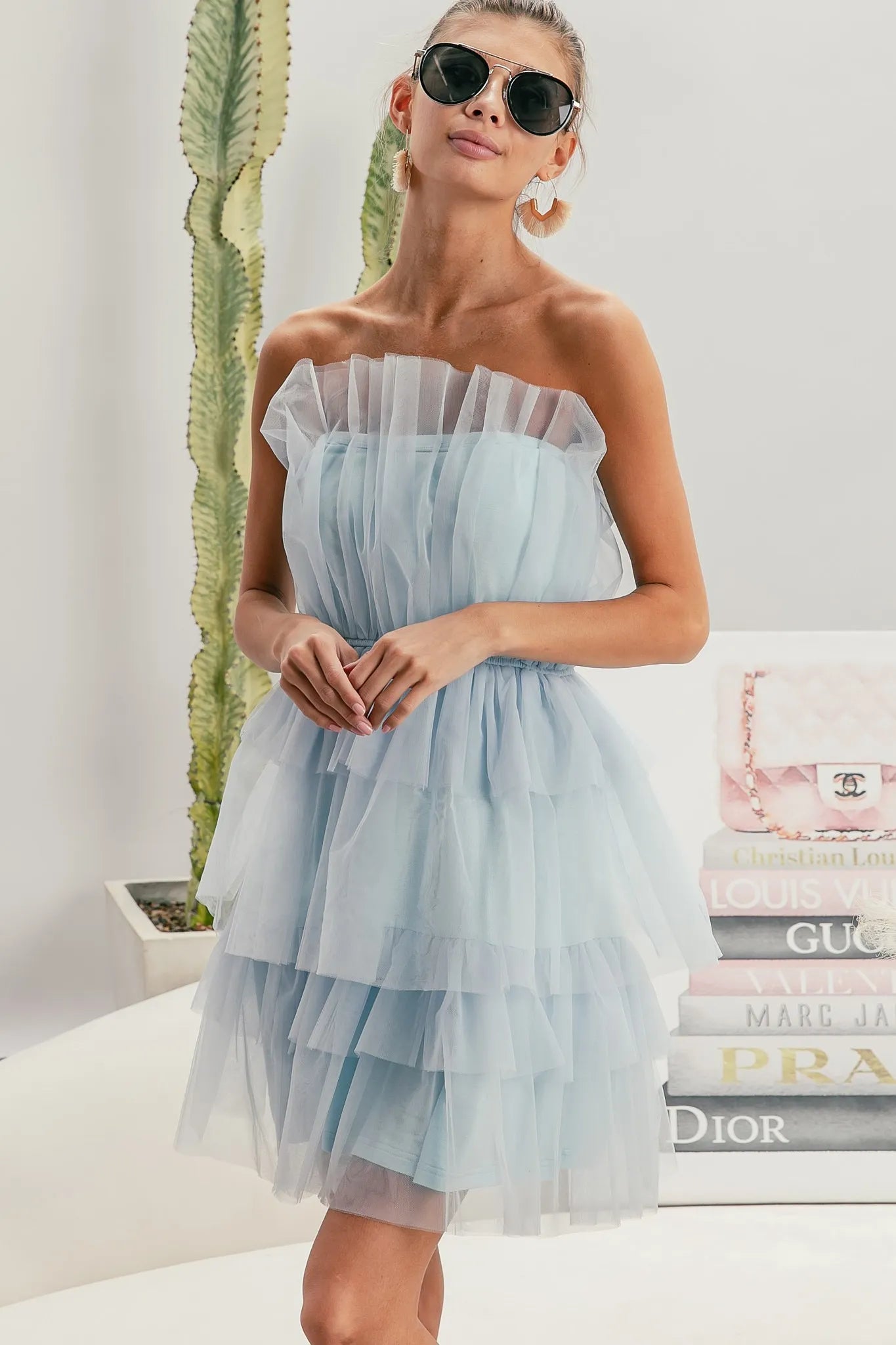 Woman wearing a light blue strapless dress with sunglasses, standing in front of a stack of fashion magazines.