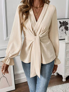 NEWYORKLISTED STYLE COLLECTION: Woman wearing a beige wrap blouse with long sleeves and tied waist, standing in a room with decor elements.