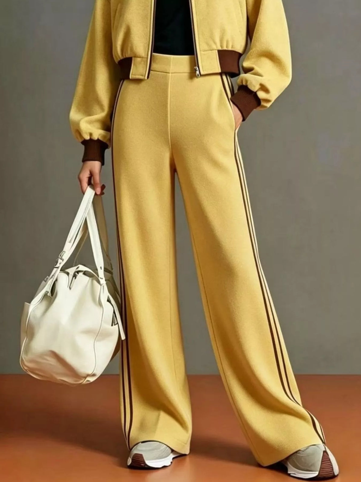 NEWYORKLISTED STYLE COLLECTION:  A model wearing a matching yellow tracksuit with brown trim, featuring wide-leg pants with double dark stripes down the side, a cropped jacket, and sneakers, while holding a white shoulder bag against a gray studio background.