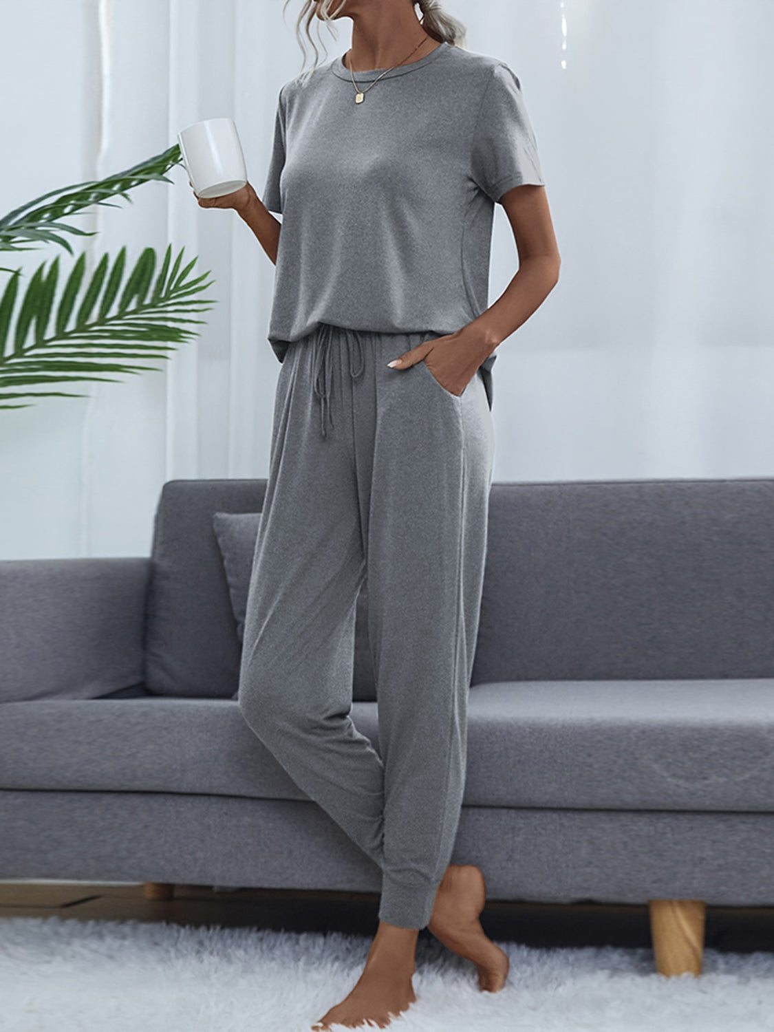 NEWYORKLISTED Intimate Apparel: Woman wearing a gray pajama set sitting on a gray couch with a white wall and plant in the background.