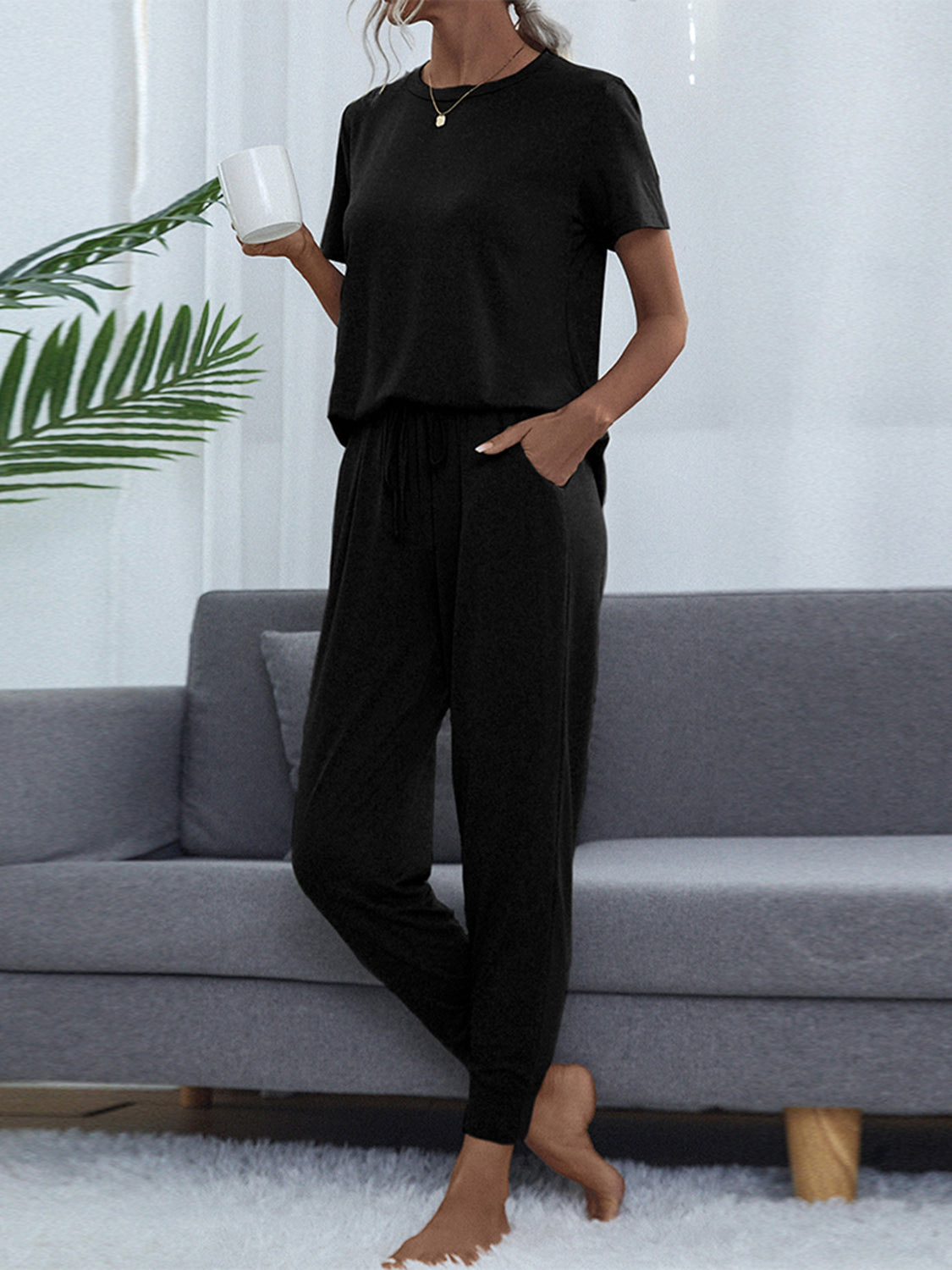 NEWYORKLISTED Intimate Apparel: Person wearing a black outfit sitting on a gray couch with a white wall and plant in the background