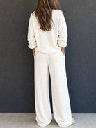 NEWYORKLISTED STYLE & COMFORT COLLECTION: A woman viewed from behind wearing a matching cream-colored knit lounge set consisting of a long-sleeve top and wide-leg pants, standing against a dark gray wall.