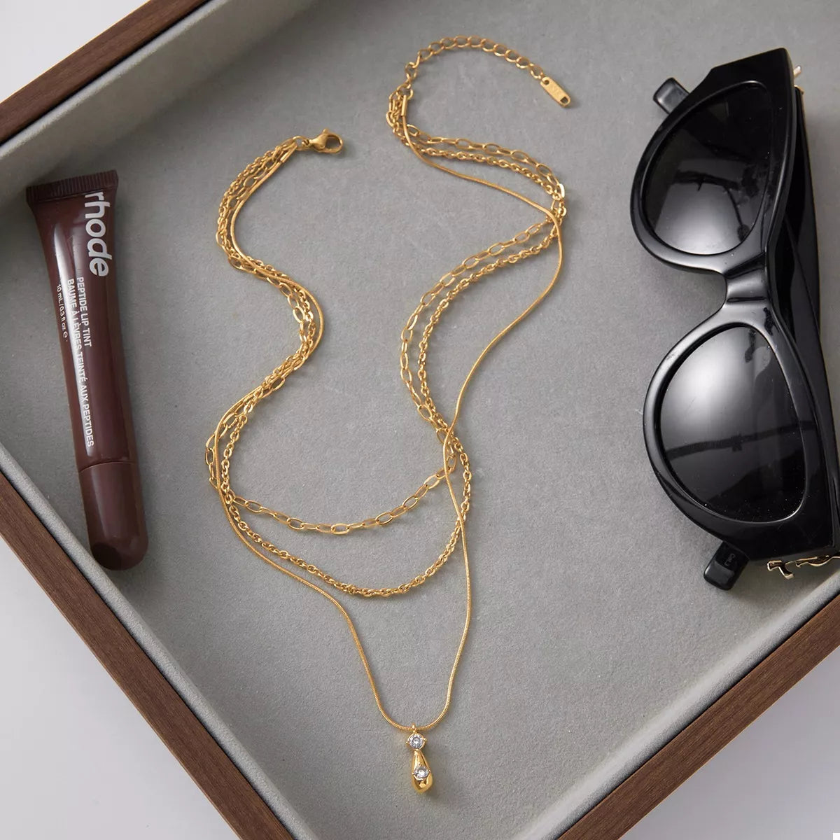 NYLD Glamour Collection by NEWYORKLISTED: Gold necklace with a pendant, tube of lipstick, and sunglasses on a gray surface.