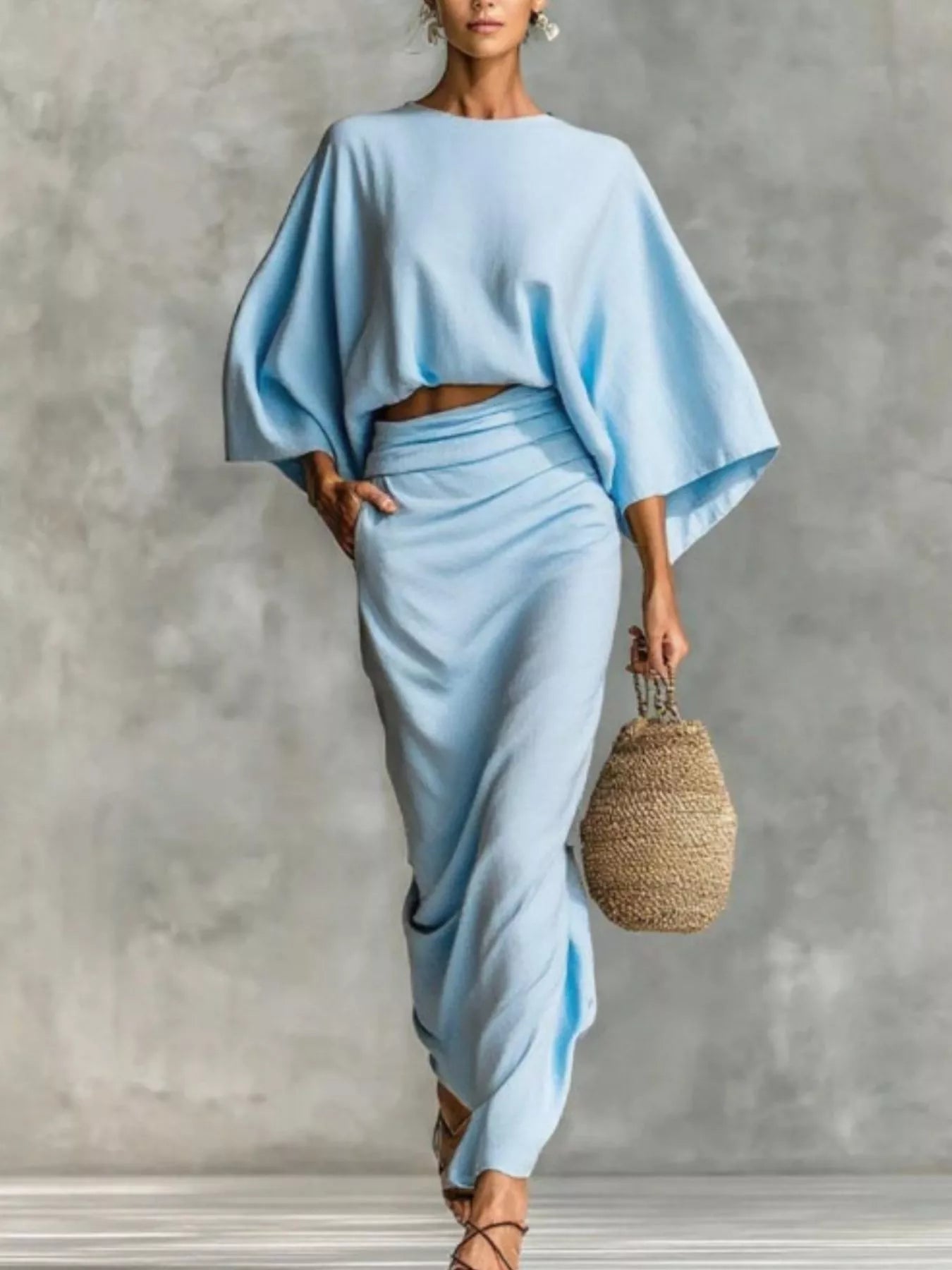 NEWYORKLISTED STYLE COLLECTION: Woman wearing a light blue outfit holding a woven bag against a gray background