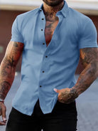 NEWYORKLISTED ATLAS MEN's COLLECTION: Man wearing a light blue button-up shirt with tattoos on arms, standing against a blurred background.
