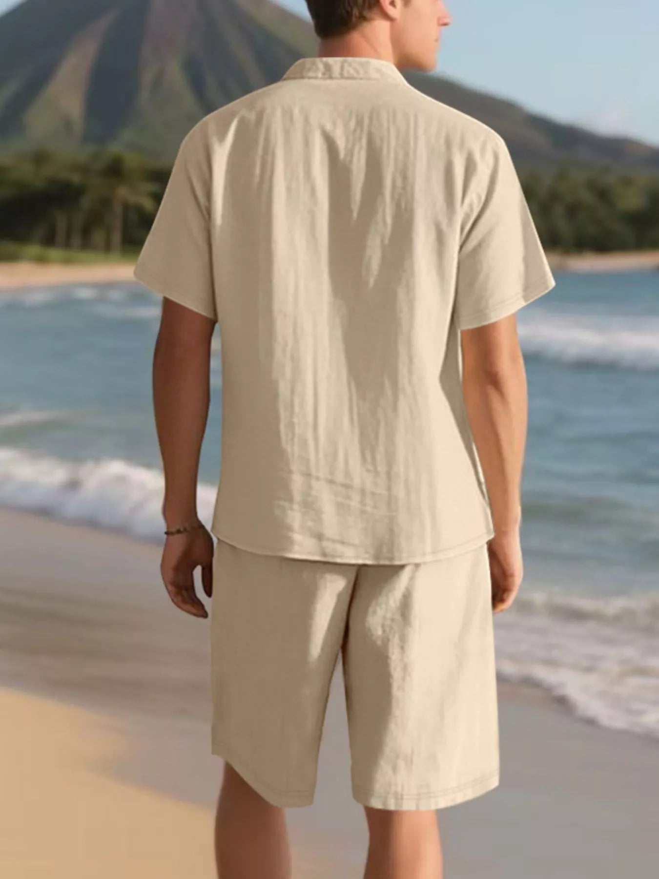 NEWYORKLISTED ATLAS MEN'S COLLECTION: Man in beige shirt and shorts standing on a beach with mountains in the background