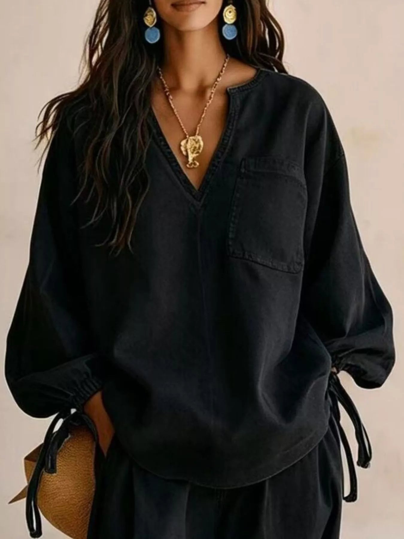 NEWYORKLISTED STYLE COLLECTION: A woman wearing an oversized black long-sleeve blouse with a split neckline, chest pocket, and side tie details, styled with gold jewelry and blue earrings against a neutral beige background.