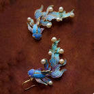 NYLD GLAMOUR By NEWYORKLISTED: Pair of blue fish earrings with gold accents and pearl-like beads on a brown background