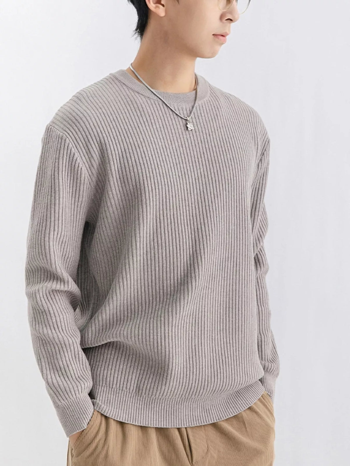 NEWYORKLISTED ATLAS MEN'S COLLECTION: A person wearing a light gray ribbed knit crewneck sweater, paired with tan pants and a silver necklace, photographed against a plain light gray background.