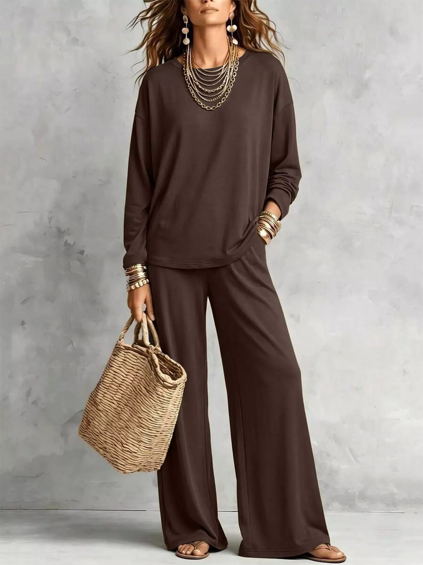 NEWYORKLISTED STYLE COLLECTION: A woman models a dark brown two-piece lounge set consisting of a long-sleeve top and matching wide-leg pants, styled with layered gold necklaces, bracelets, and a straw tote bag against a textured gray wall.