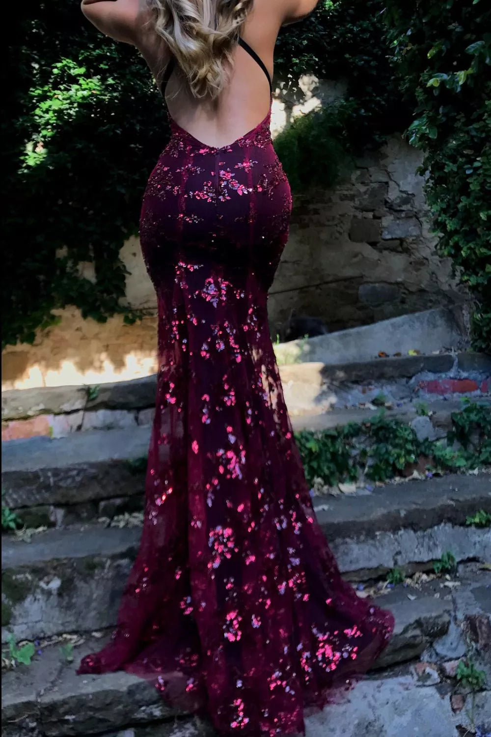 Person wearing a sparkly burgundy dress on stone steps with greenery in the background