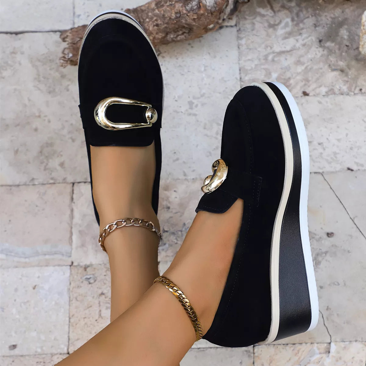 NEWYORKLISTED SHOE COLLECTION: Black slip-on shoes with gold accents worn by a person on a light stone floor.
