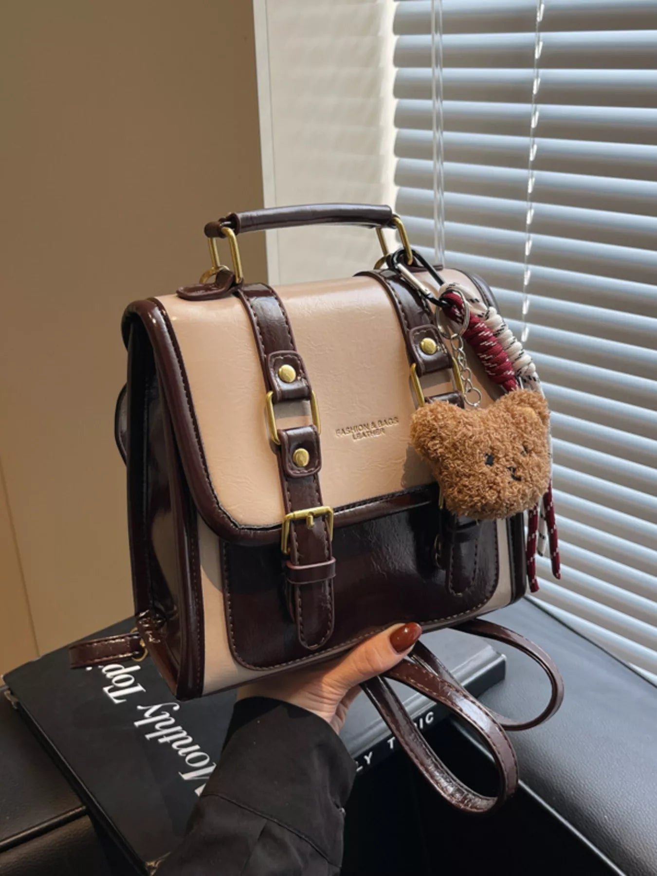 NEWYORKLISTED STYLE COLLECTION: Beige and brown handbag with teddy bear charm held by a person, with a window and blinds in the background.