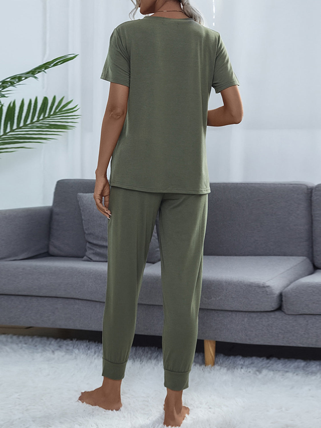 NEWYORKLISTED Intimate Apparel: Person wearing olive green pajamas standing in a living room with a gray sofa and white walls.